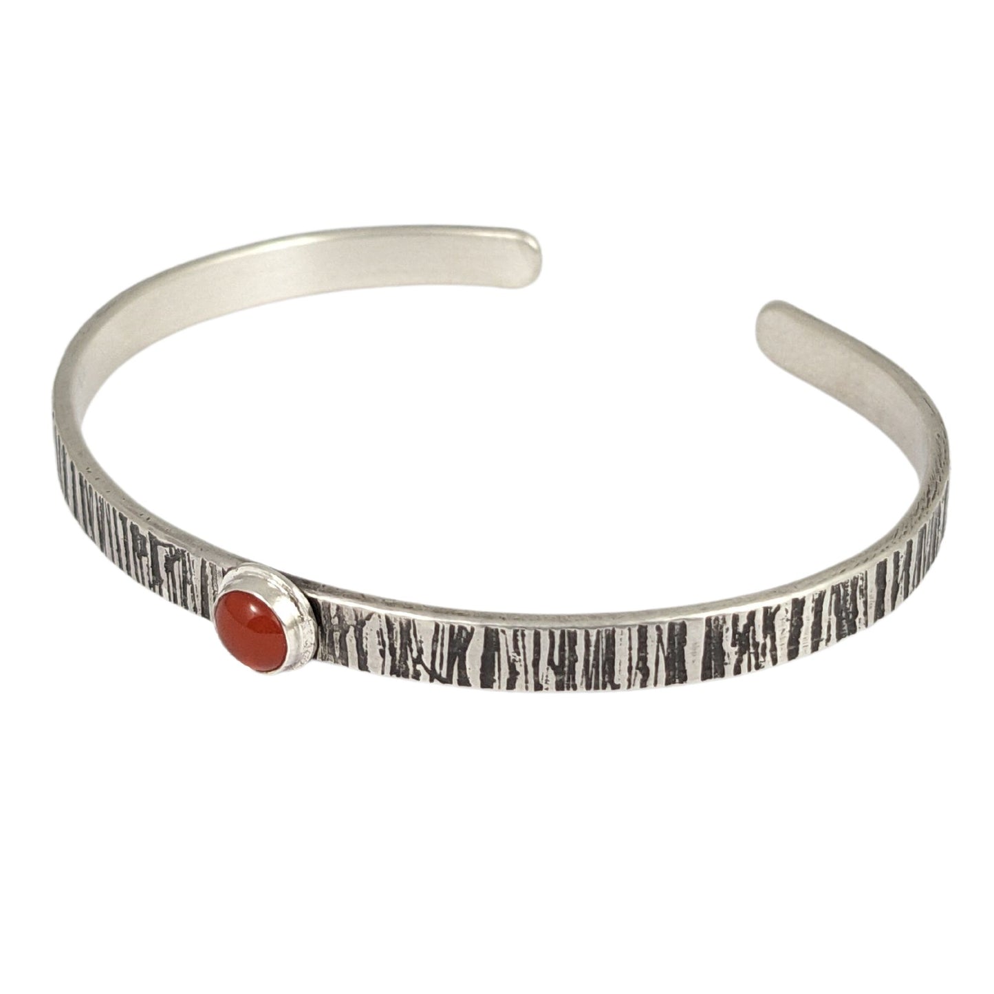 Sterling silver cuff bracelet. The narrow rectangular bracelet has a random lines texture that is oxidized to darken the design. In the middle is an orange carnelian gemstone. The gemstone is round on top.