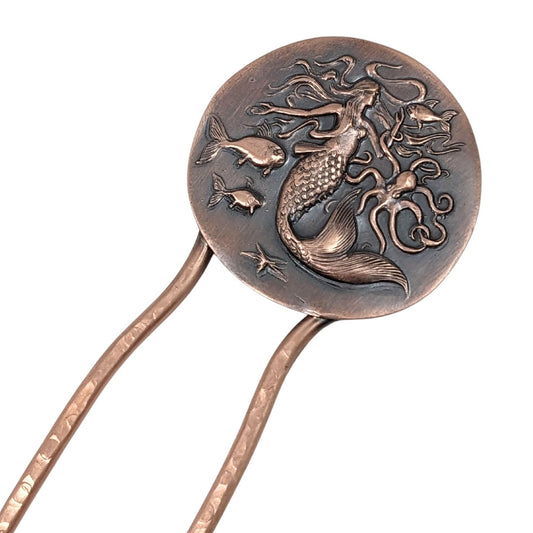 Copper hair fork. At the top is a large round disk with a sea life design impressed in the copper. There is a large mermaid with flowing hair in the center, an octopus to her left, three fish, and a starfish.