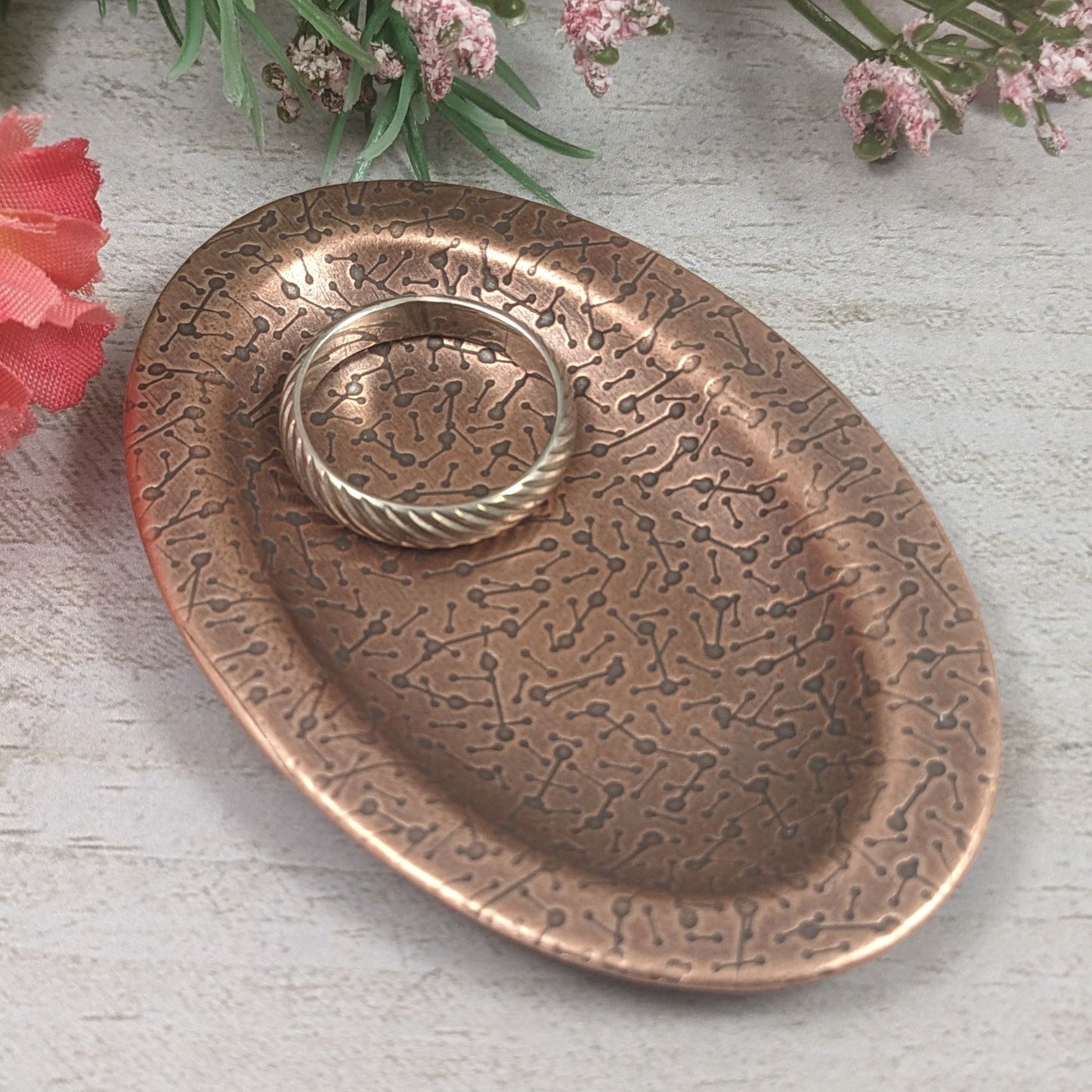 Oval ring dish made of copper. The dish has a shallow bowl with a lip around the edge. It is covered in a pattern meant to represent a meteor shower. The pattern is impressed into the copper and is short lines with small solid circles at the end. The design is oxidized, which means the impressed parts are dark, almost black. Staged with flowers in the background and a ring in the dish.