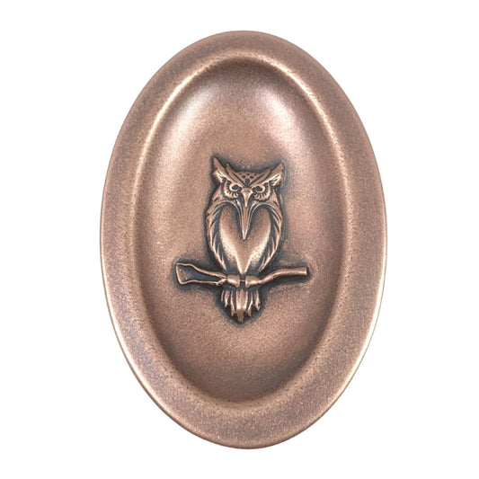 Oval ring dish made of copper. The dish has a shallow bowl with a lip around the edge. Centered on the bottom of the dish is a three-dimensional design of an owl, also made of copper. The owl is facing straight ahead and is perched on a branch. The breast is smooth and shaped like a long narrow heart. The owl hd large eyes, a pointed beak, tufted ears, and feathers around the breast and on the tail.  The design is oxidized, which means the recessed parts of the owl are dark, almost black.