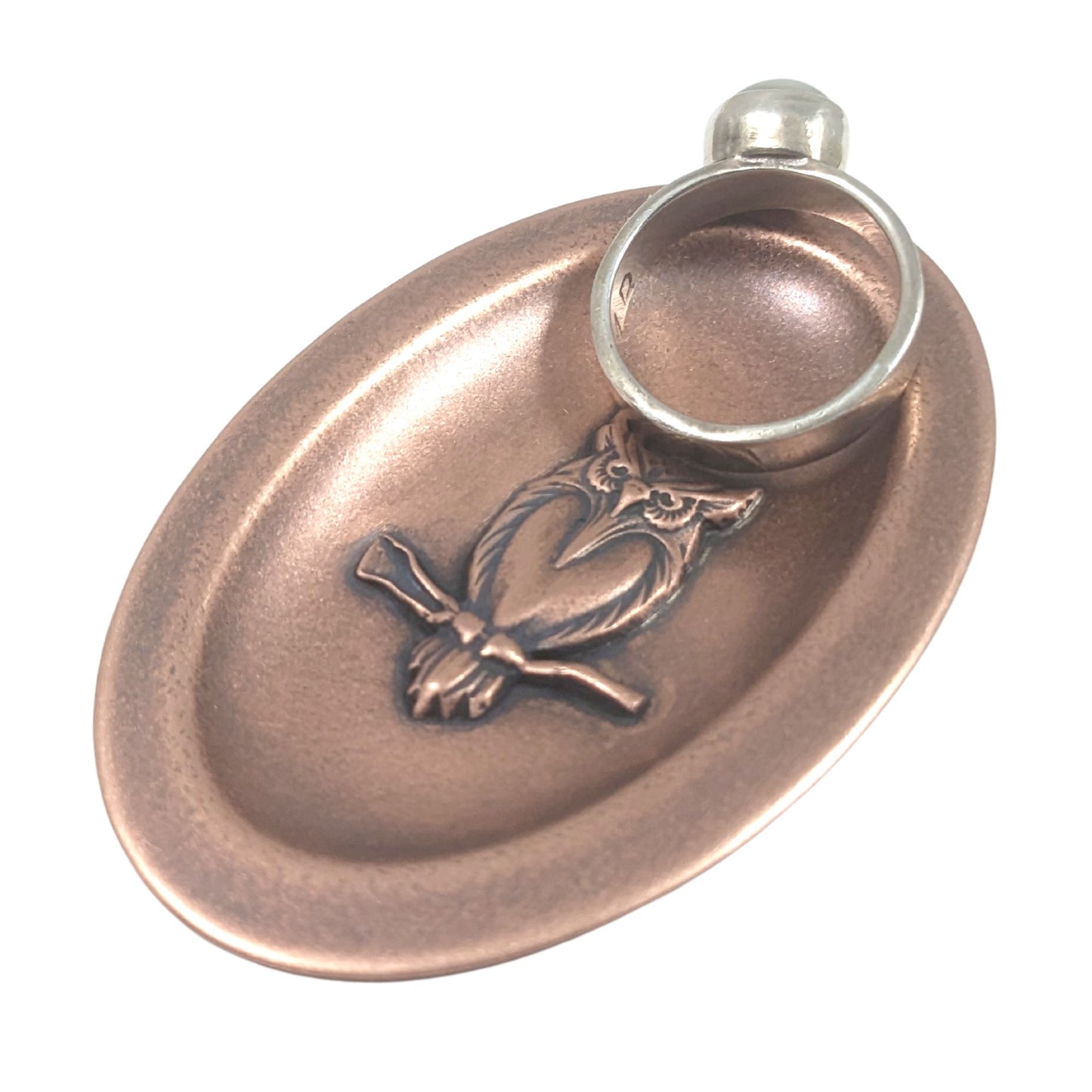 Oval ring dish made of copper. The dish has a shallow bowl with a lip around the edge. Centered on the bottom of the dish is a three-dimensional design of an owl, also made of copper. The owl is facing straight ahead and is perched on a branch. The breast is smooth and shaped like a long narrow heart. The owl hd large eyes, a pointed beak, tufted ears, and feathers around the breast and on the tail. Staged with a ring.