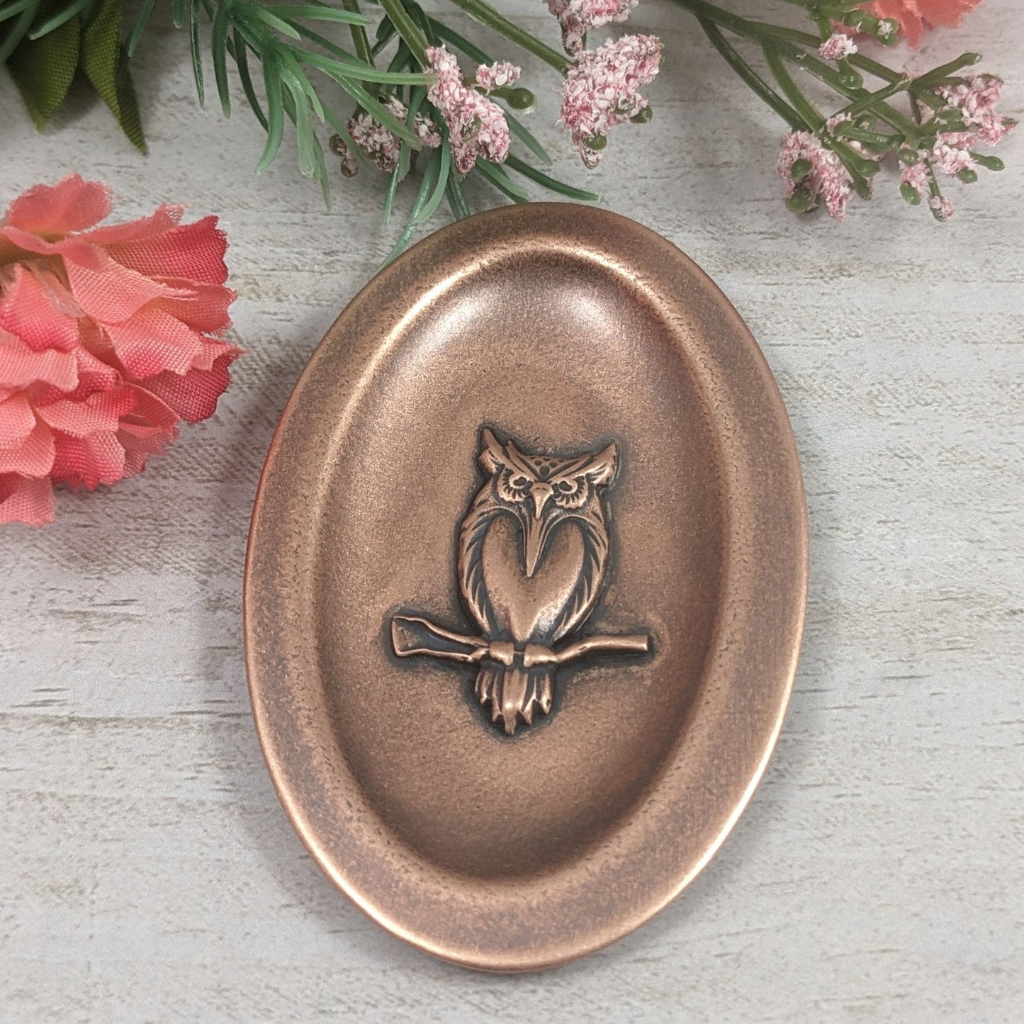 Oval ring dish made of copper. The dish has a shallow bowl with a lip around the edge. Centered on the bottom of the dish is a three-dimensional design of an owl, also made of copper. The owl is facing straight ahead and is perched on a branch. The breast is smooth and shaped like a long narrow heart. The owl hd large eyes, a pointed beak, tufted ears, and feathers around the breast and on the tail.  Staged with flowers