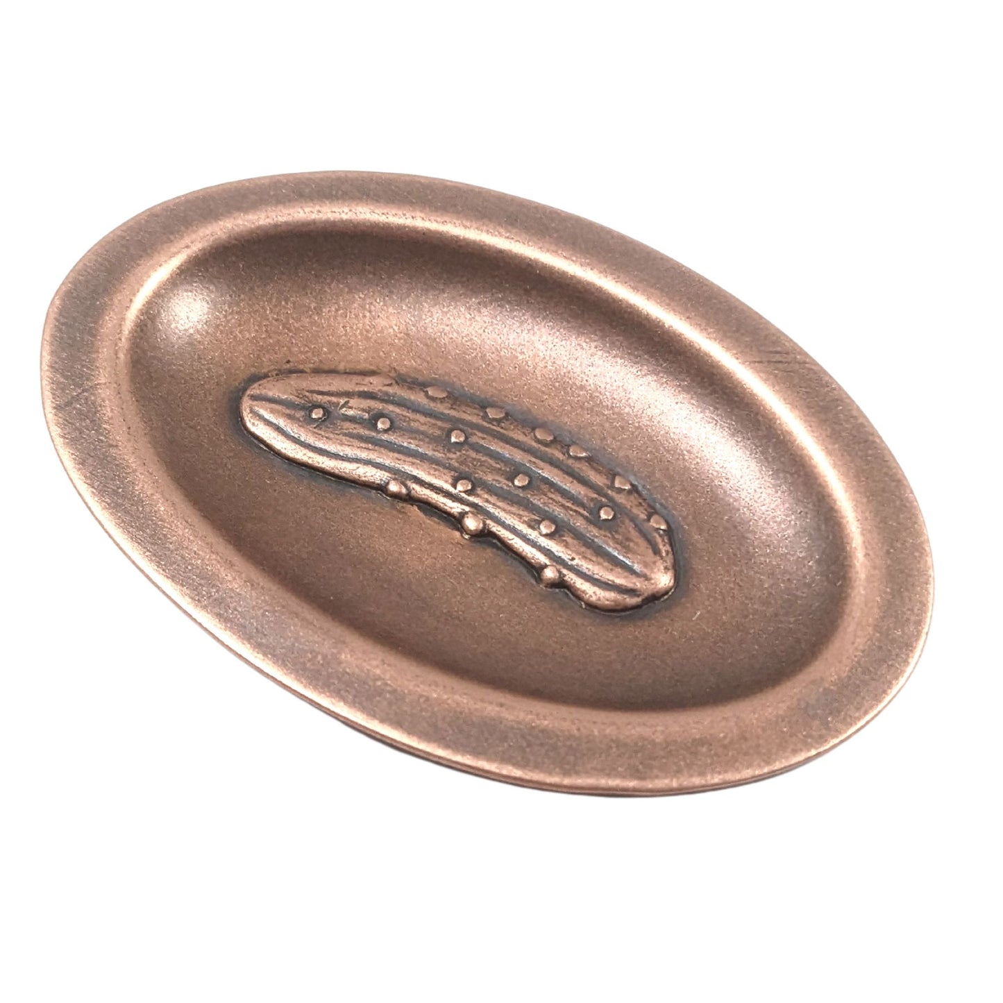 Oval ring dish made of copper. The dish has a shallow bowl with a lip around the edge. Centered on the bottom of the dish is a three-dimensional design of a pickle, also made of copper. The pickle is large and has stipples and ridges, just like a fresh from the garden pickle would have. The design is oxidized, which means the recessed parts of the pickle are dark, almost black.