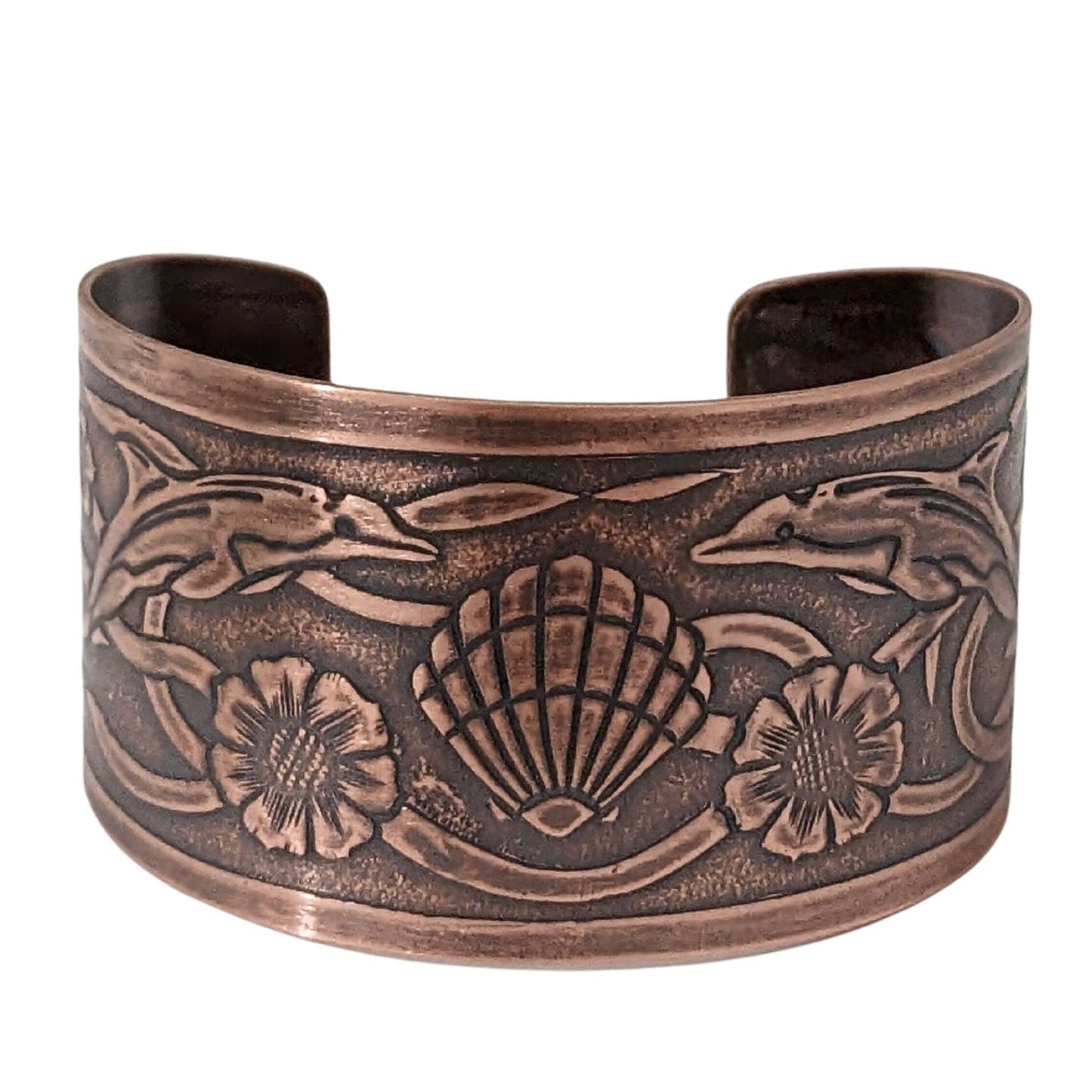 Wide tapered copper cuff bracelet with sea life design. Center is a seashell, with flowers and dolphins on either side. Toward the ends of the cuff there are turtles, and a seaweed pattern is woven throughout the design.