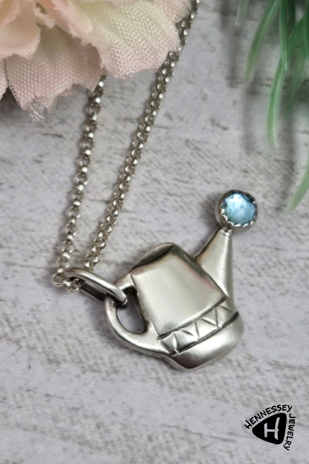 Sterling silver watering can pendant with a blue topaz gemstone at the end of the spout to represent water.