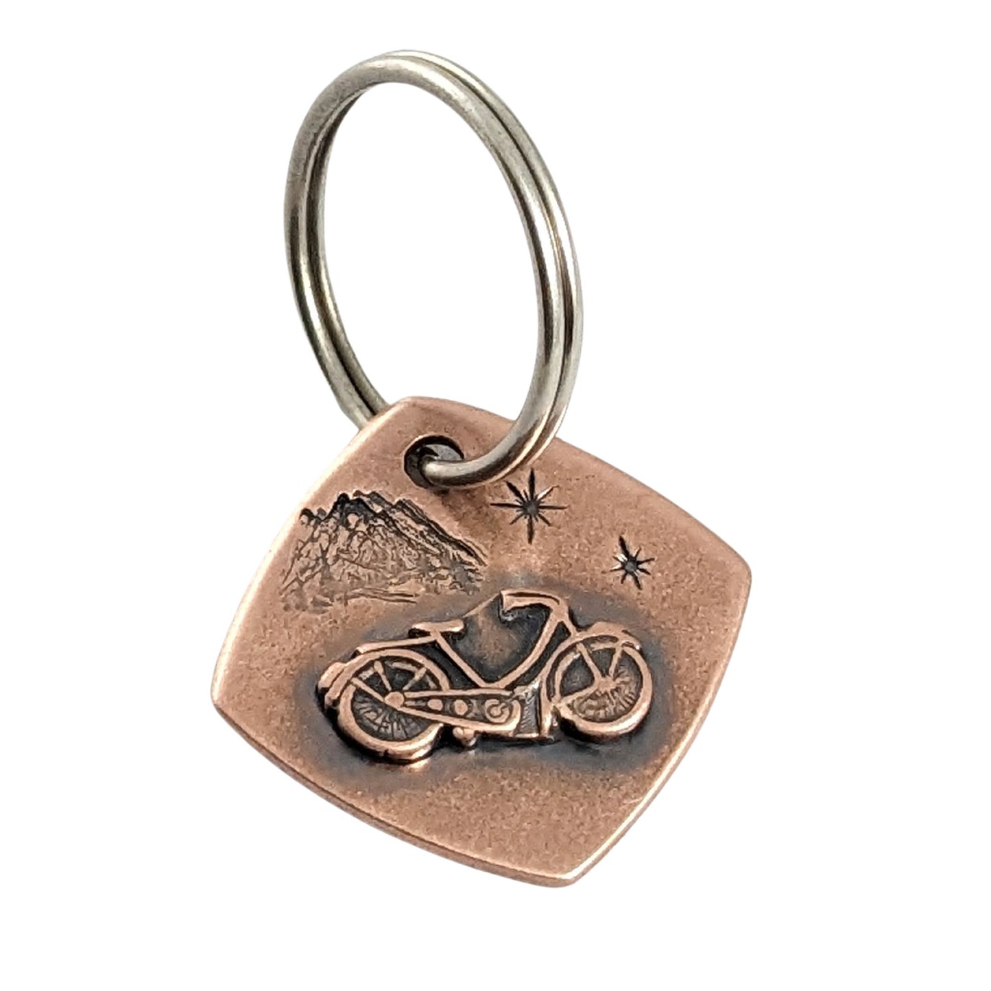 copper keychain with raised detailed bicycle. background is mountains and twinkling stars