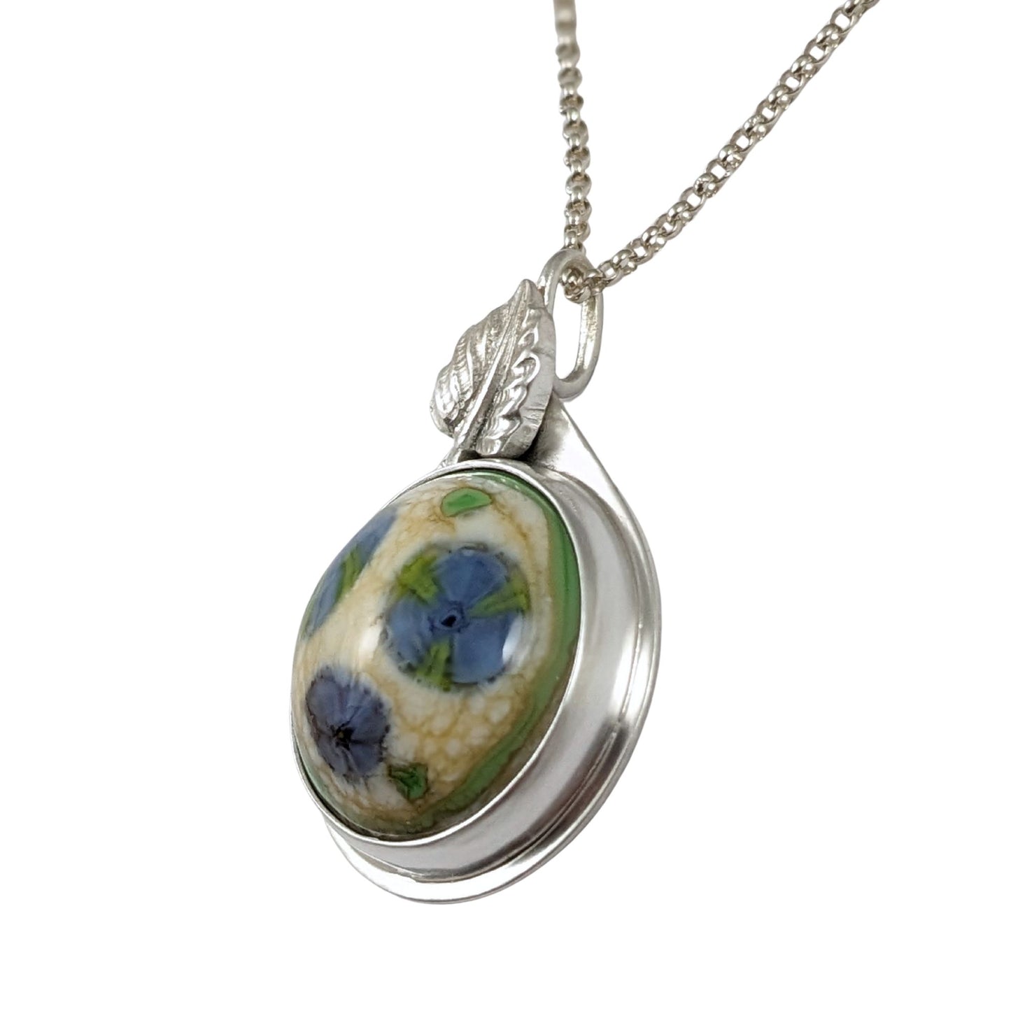 Sterling silver pendant. The stone is a round lampwork glass cabochon with three blue flowers. The flowers have bits of green in them. The background is pale yellow and there is a border of green. Above the stone and partially hiding the bale is a silver leaf.