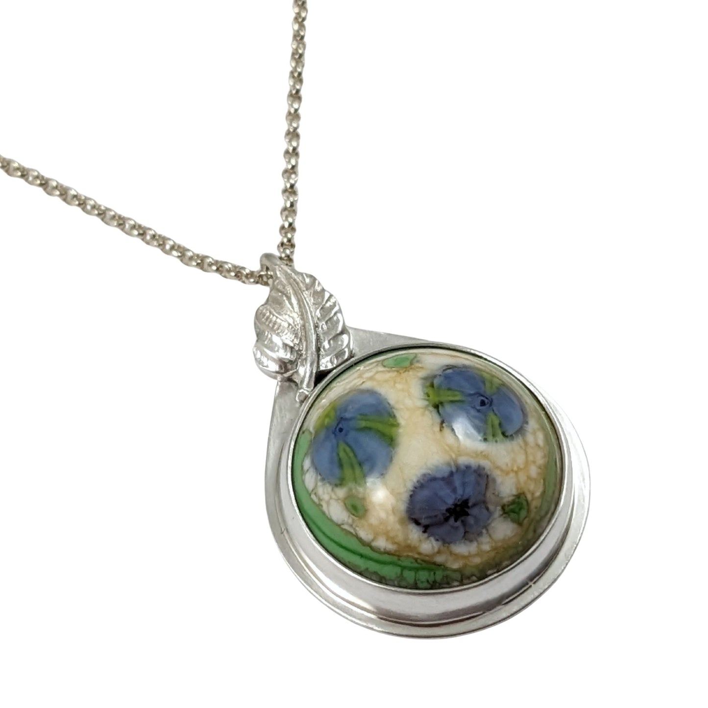 Sterling silver pendant. The stone is a round lampwork glass cabochon with three blue flowers. The flowers have bits of green in them. The background is pale yellow and there is a border of green. Above the stone and partially hiding the bale is a silver leaf.