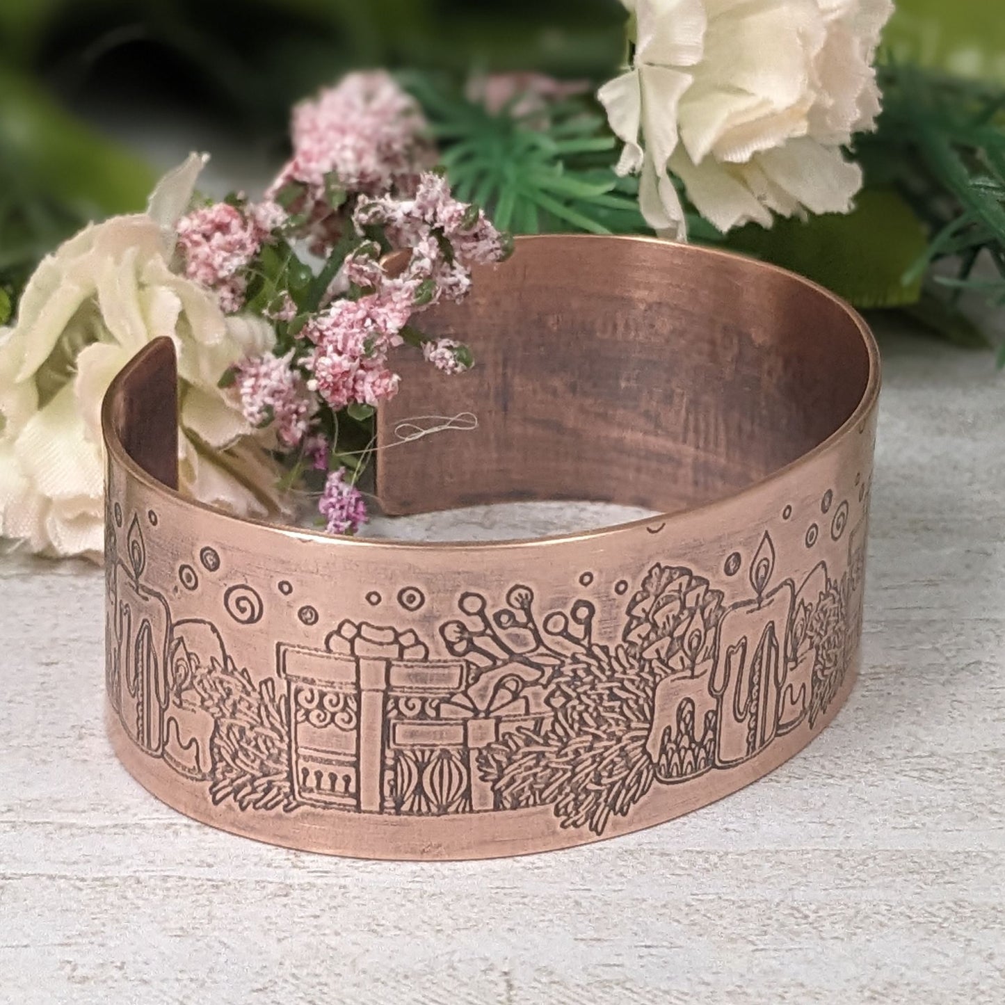 copper cuff bracelet with a christmas theme. the cuff is covered in impressions of candles, wrapped gifts, and evergreens