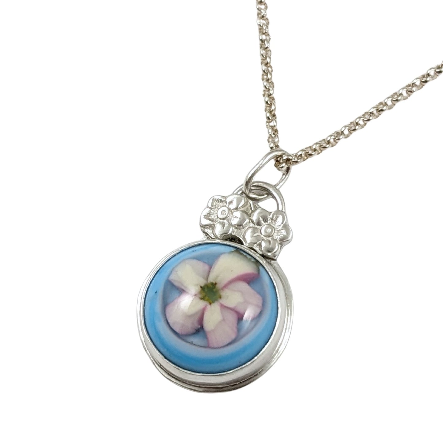 Sterling silver pendant with a glass cabochon. The cabochon has a white five petal flower and a blue background. Above the cabochon setting there are two small five petal flowers made from sterling silver, then a small bail.