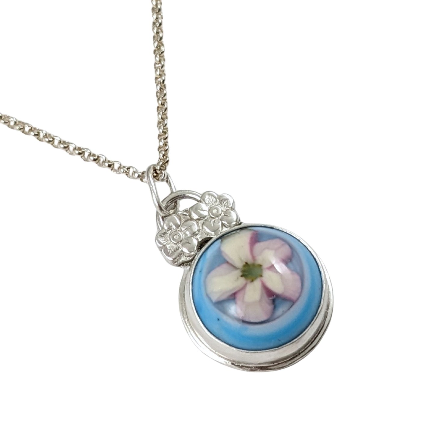 Sterling silver pendant with a glass cabochon. The cabochon has a white five petal flower and a blue background. Above the cabochon setting there are two small five petal flowers made from sterling silver, then a small bail.