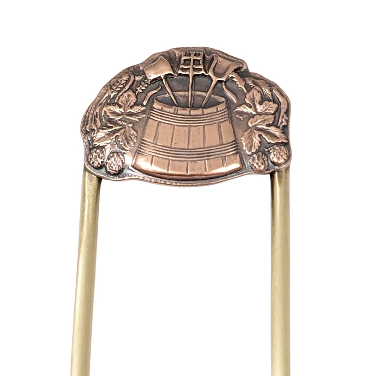 brass hair fork with a dimensional design in copper. The design has a wooden barrel in the middle, filled with three long handled garden tools. Surrounding the barrel are leaves and strawberries.