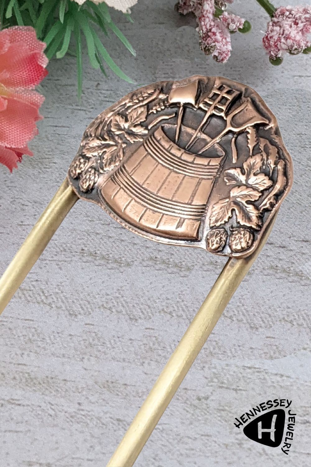 brass hair fork with a dimensional design in copper. The design has a wooden barrel in the middle, filled with three long handled garden tools. Surrounding the barrel are leaves and strawberries.