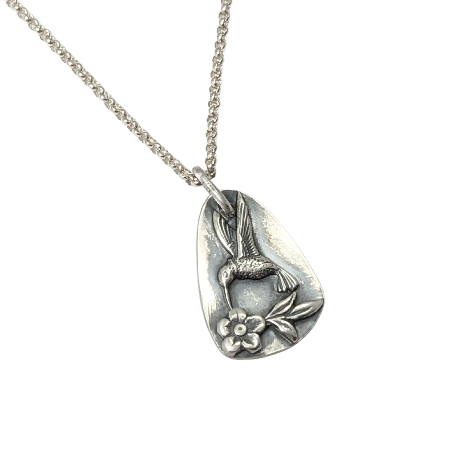 Sterling silver pendant in a freeform oval shape. On the pendant there is a silver hummingbird in flight, hovering over a single flower with three leaves.