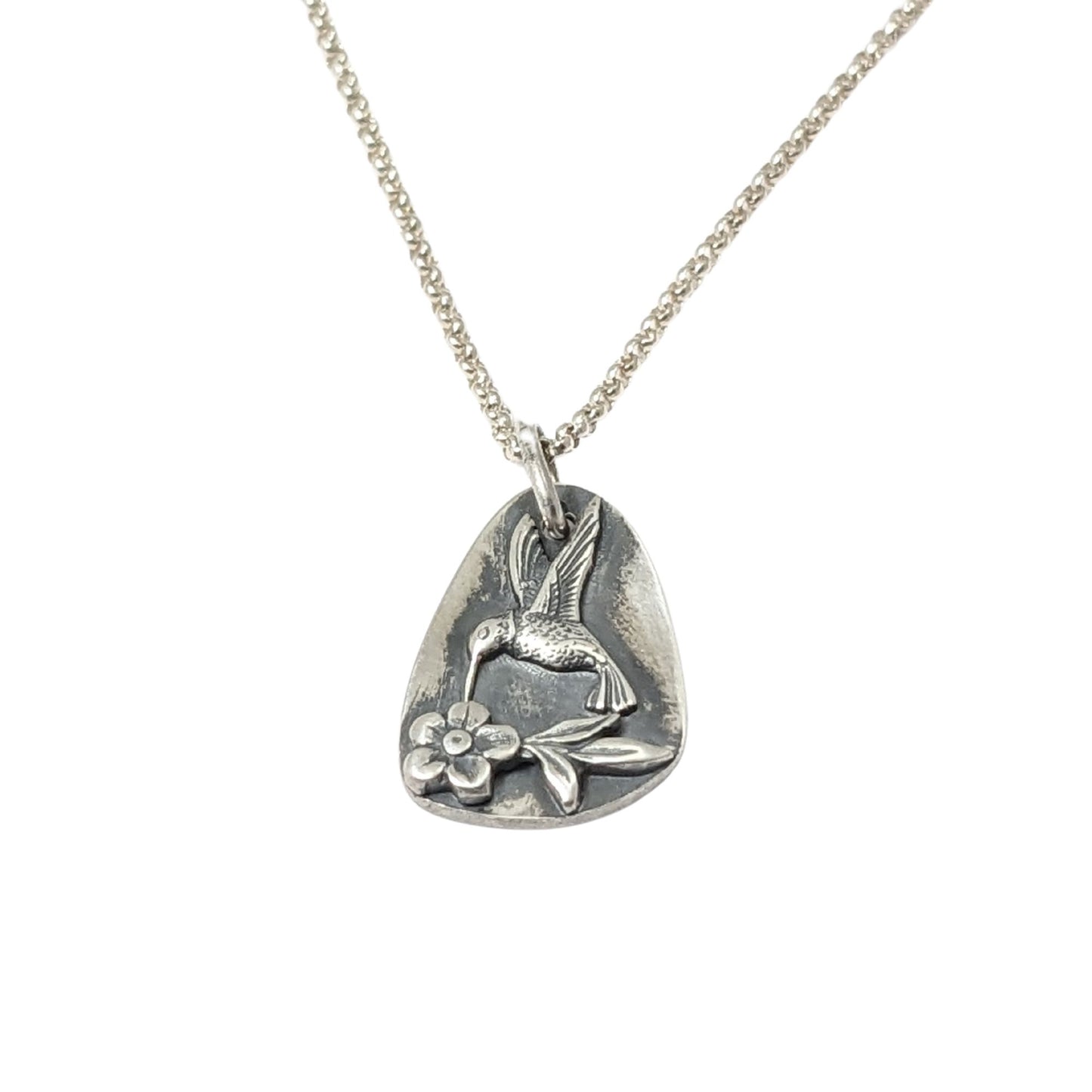 Sterling silver pendant in a freeform oval shape. On the pendant there is a silver hummingbird in flight, hovering over a single flower with three leaves.