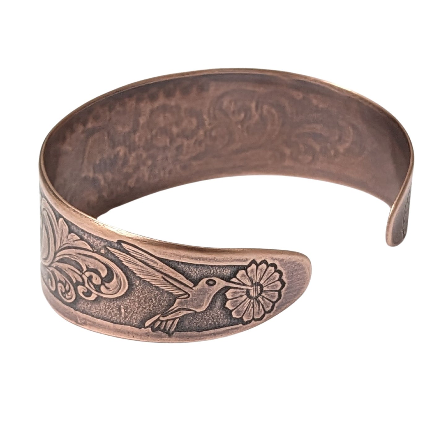 Tapered copper cuff bracelet. Pattern across middle is a hummingbird surrounded by flowers. At each end of the cuff there is a smaller hummingbird and flower.