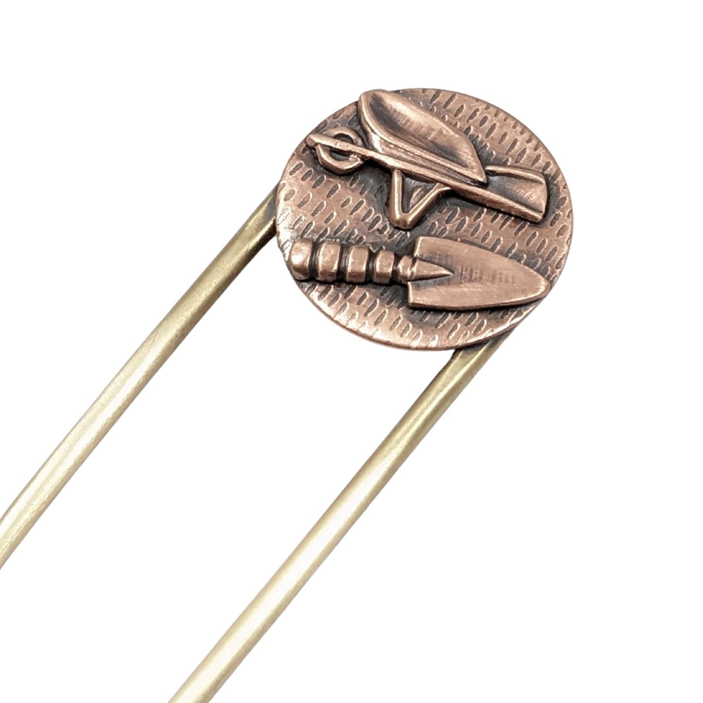 Copper hair fork with a round design on the end of the fork. The design is a round piece of copper textured to resemble seeds. Soldered to the round is a copper wheelbarrow and a copper hand trowel. The fork is brass.