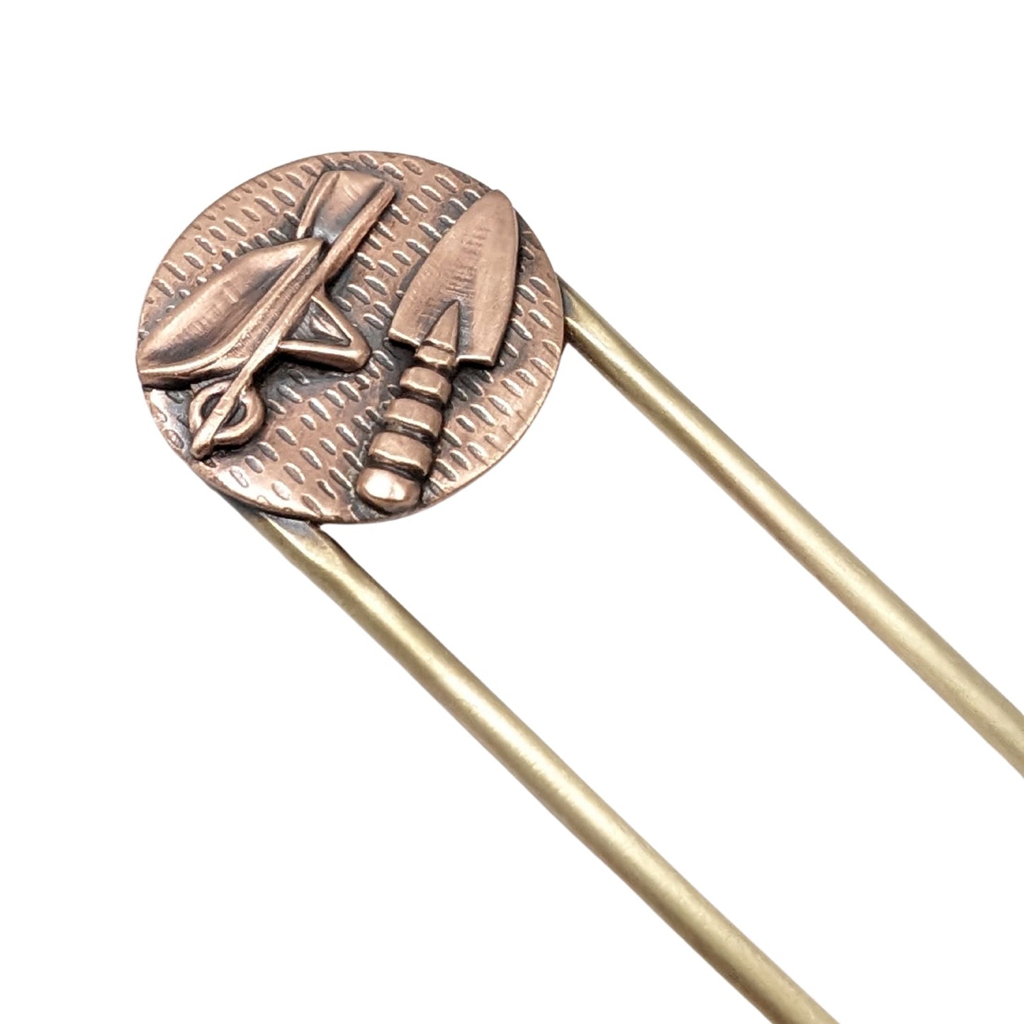 Copper hair fork with a round design on the end of the fork. The design is a round piece of copper textured to resemble seeds. Soldered to the round is a copper wheelbarrow and a copper hand trowel. The fork is brass.