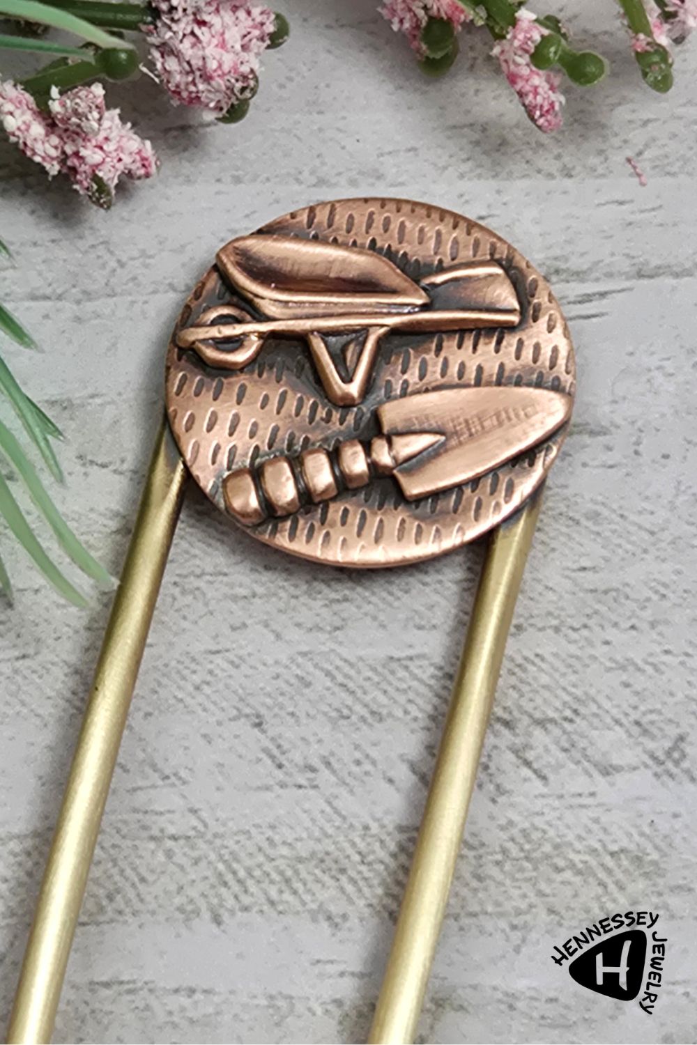 Copper hair fork with a round design on the end of the fork. The design is a round piece of copper textured to resemble seeds. Soldered to the round is a copper wheelbarrow and a copper hand trowel. The fork is brass.