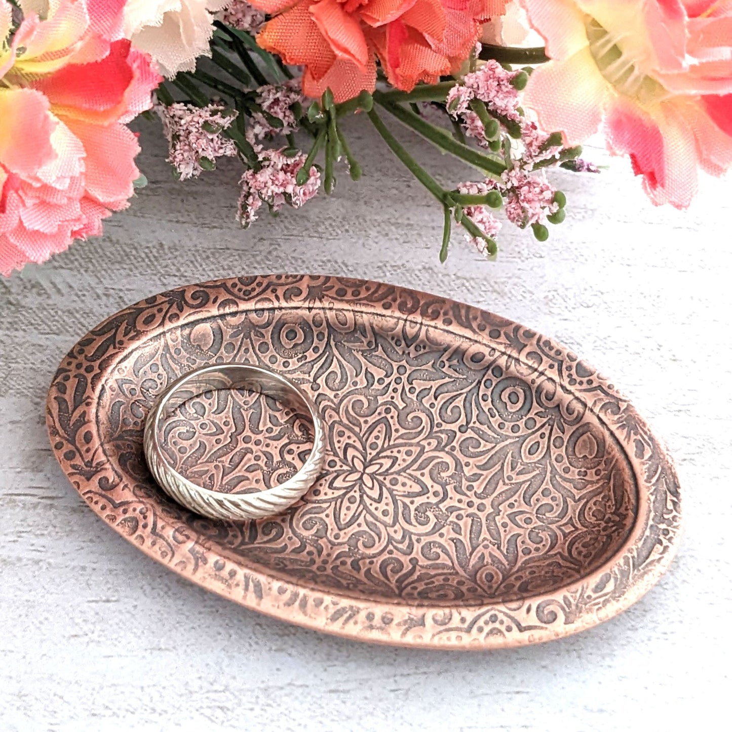 Copper oval ring dish with abstract garden flowers and leaves design. Dish is 2 inches by three inches with a raised lip. displayed with flowers in background