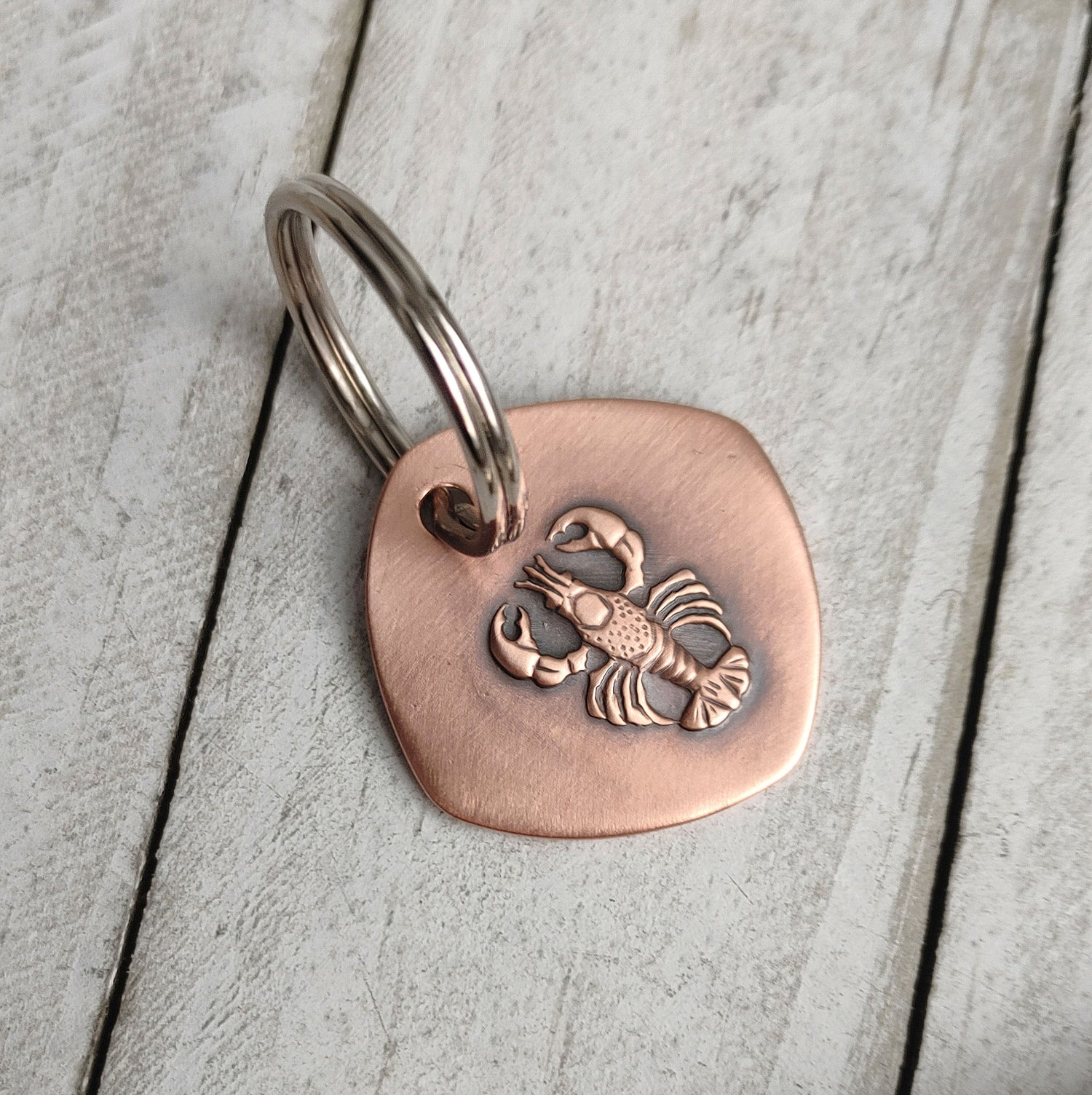 Copper keychain with a detailed raised impression of a lobster. The claws, legs, and tail are all clearly defined. The keychain is a rounded square shape.