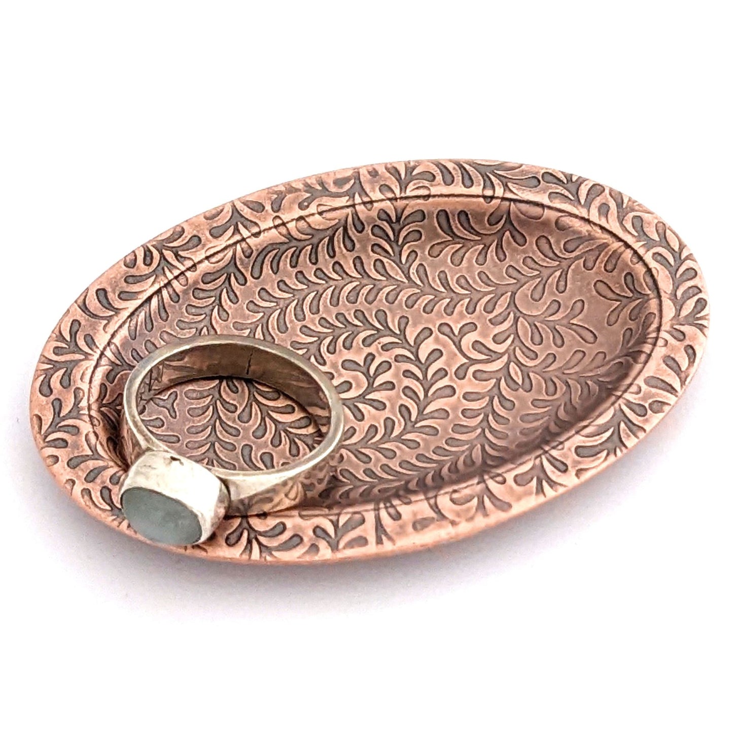 Copper oval ring dish with abstract fiddlehead fern pattern. Dish is 2 inches by three inches with a raised lip.