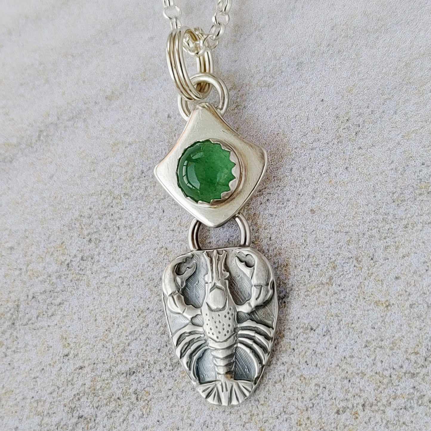 This pendant is a finely detailed raised lobster impression, the gemstone is a 6mm aventurine cabochon set on a curved square background, and the round background has a hammered line texture. Aventurine is a shimmering green form of quartz. This pendant is 1-1/4 inches long and 1/2 inch across. It comes on an 18 inch sterling silver chain.