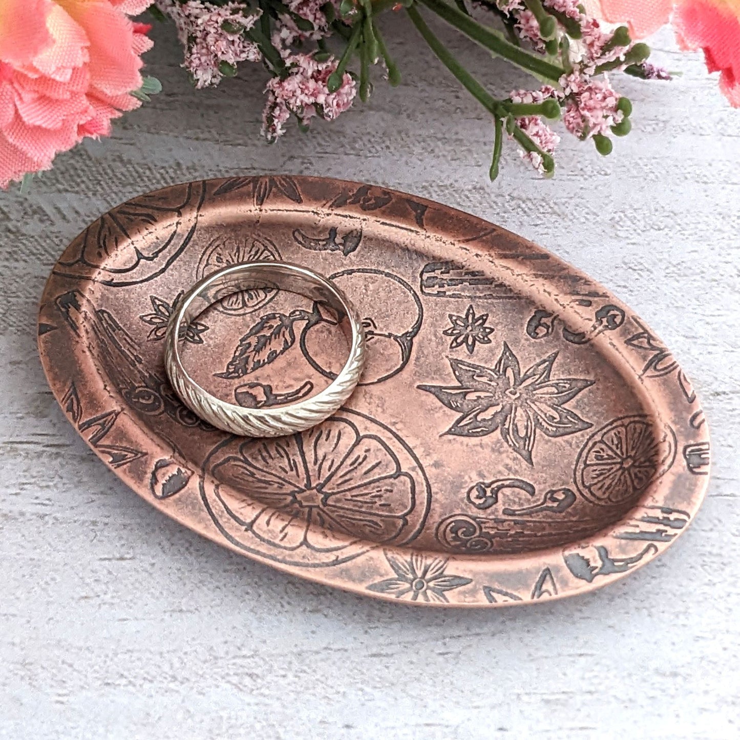 Copper oval ring dish with design of ingredients used in winter spice drinks, including apples oranges nutmeg cinnamon and clove. Dish is 2 inches by three inches with a raised lip.