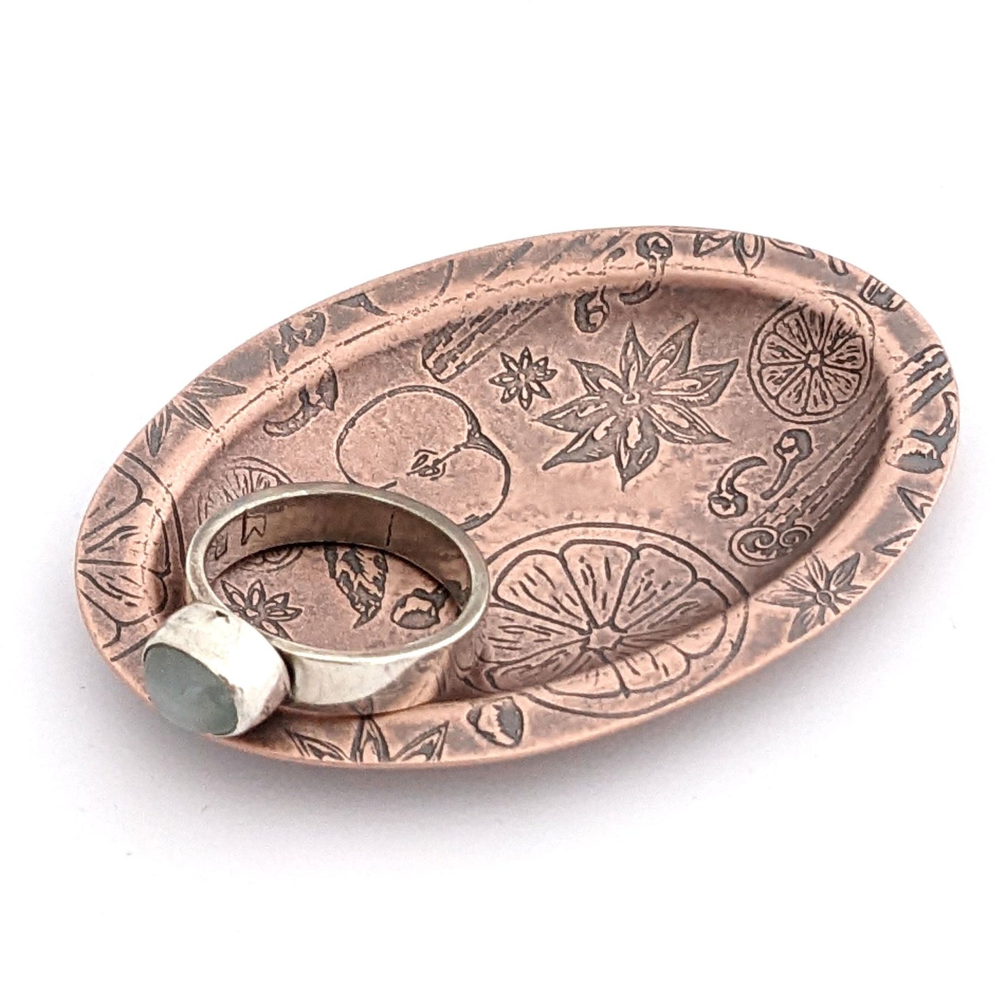 Copper oval ring dish with design of ingredients used in winter spice drinks, including apples oranges nutmeg cinnamon and clove. Dish is 2 inches by three inches with a raised lip.