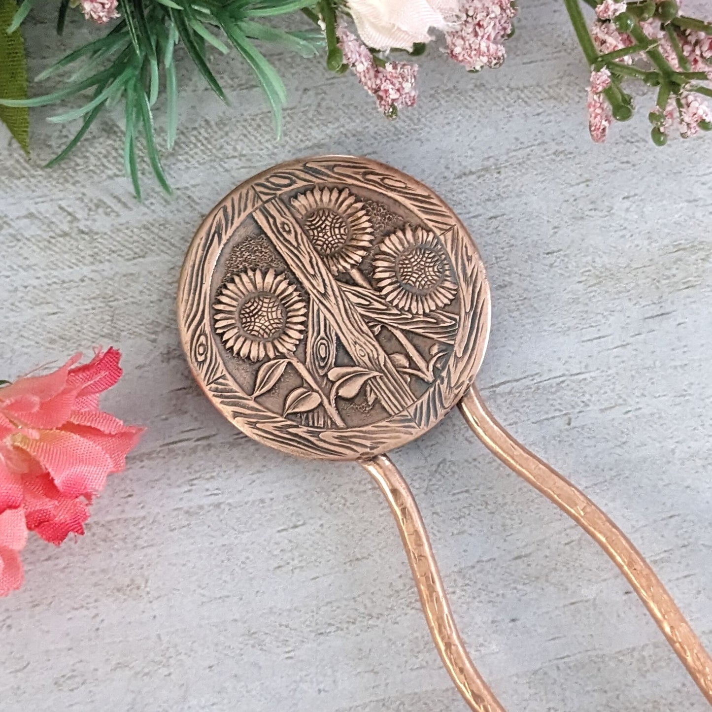 Copper hair fork with a large round design at top of sunflowers and a peace sign. The peace sign has a wood texture. Three sunflowers grow through the wood sign. There's a lot of detail - veins on the leaves, you can see the sunflower seeds and petals. On a copper fork with a hammered finish. Staged on a gray background surrounded by flowers.