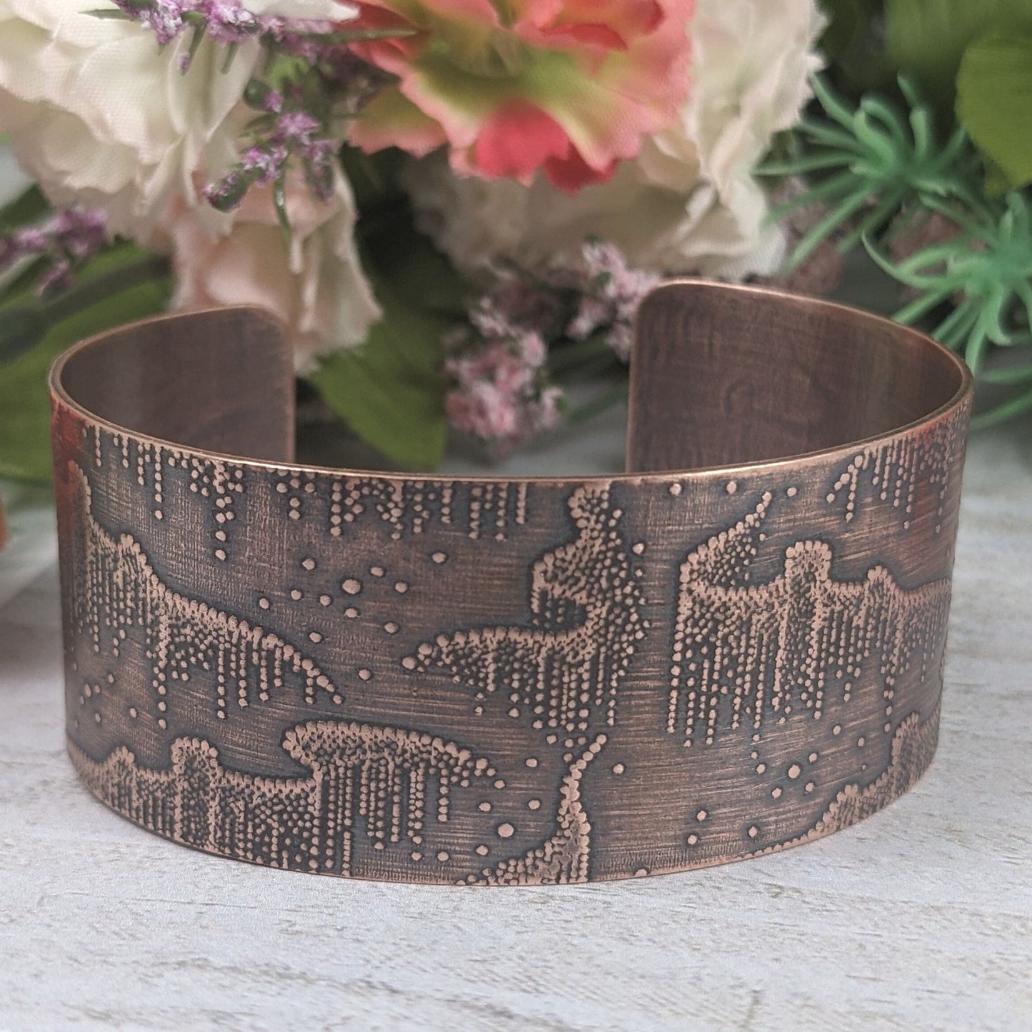 Wide copper cuff bracelet with repeated design pattern representing the aurora borealis Staged with flowers in the background