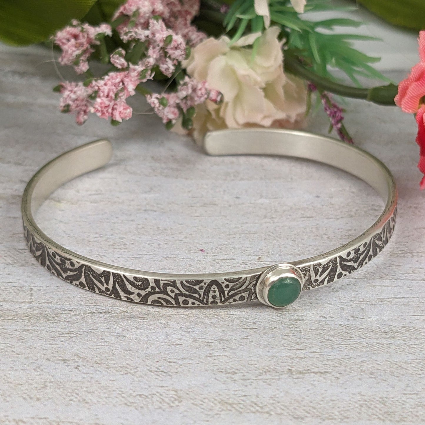 Narrow sterling silver cuff bracelet with a green aventurine gemstone. The rectangular silver wire has a n abstract impressed design, and the impression is oxidized black to bring out the details. Staged with flowers in the background.