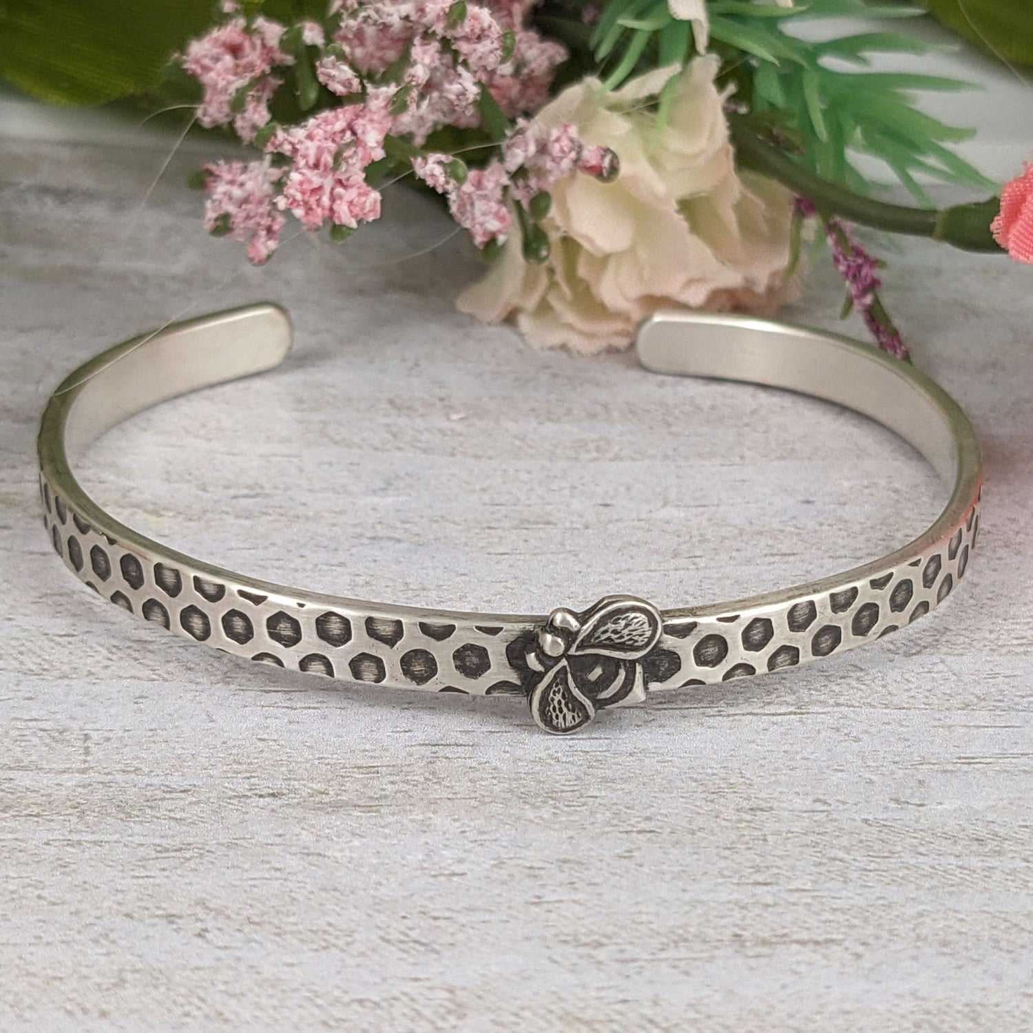 Narrow sterling silver cuff bracelet with a hexagon honeycomb pattern. A the center of the cuff there's a sterling silver honeybee. Staged with flowers in the background.