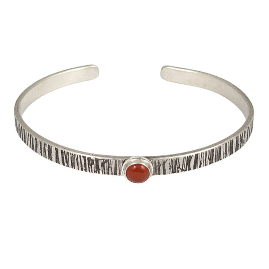 Sterling silver cuff bracelet. The narrow rectangular bracelet has a random lines texture that is oxidized to darken the design. In the middle is an orange carnelian gemstone. The gemstone is round on top.