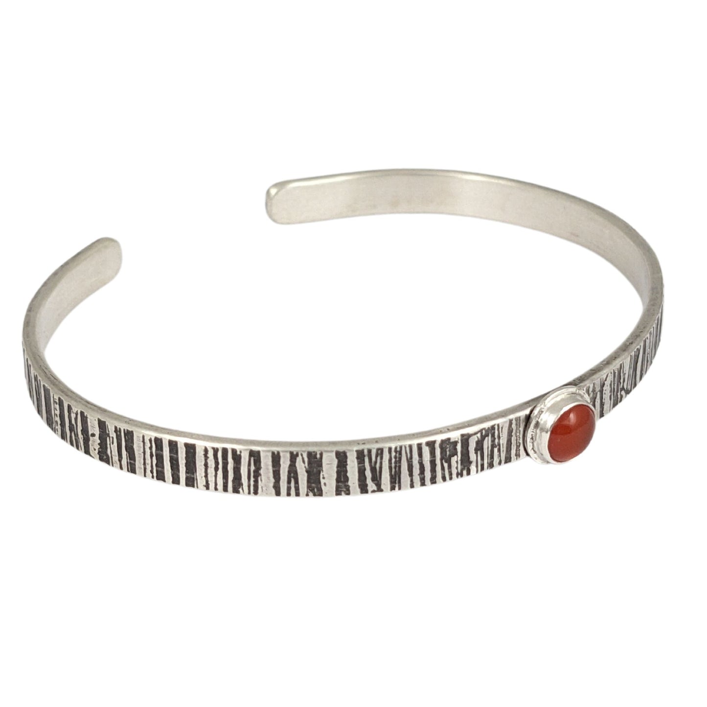 Sterling silver cuff bracelet. The narrow rectangular bracelet has a random lines texture that is oxidized to darken the design. In the middle is an orange carnelian gemstone. The gemstone is round on top.
