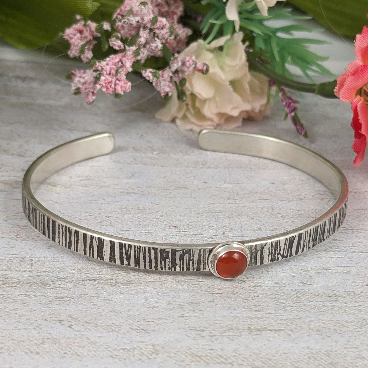 Sterling silver cuff bracelet. The narrow rectangular bracelet has a random lines texture that is oxidized to darken the design. In the middle is an orange carnelian gemstone. The gemstone is round on top. Staged with flowers in the background.