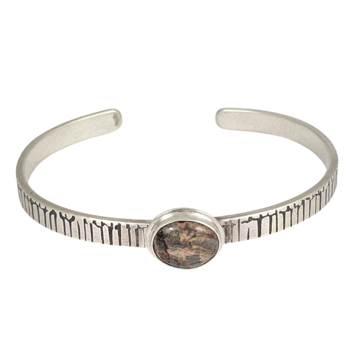 A stone in muted shades of pink, gray, and brown set on a narrow rectangular shaped sterling silver cuff bracelet. The cuff has a design that looks like an earthquake seismograph chart.