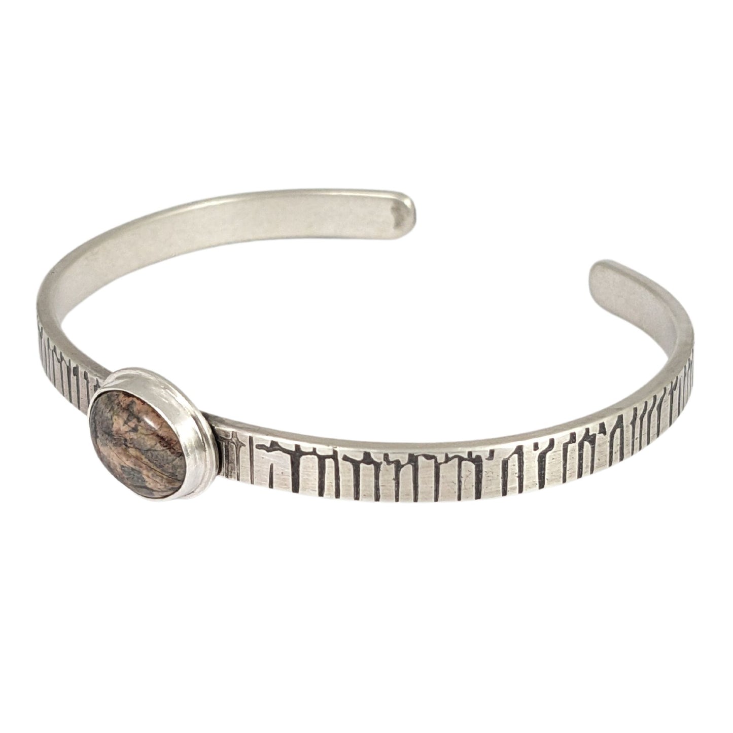 A stone in muted shades of pink, gray, and brown set on a narrow rectangular shaped sterling silver cuff bracelet. The cuff has a design that looks like an earthquake seismograph chart.