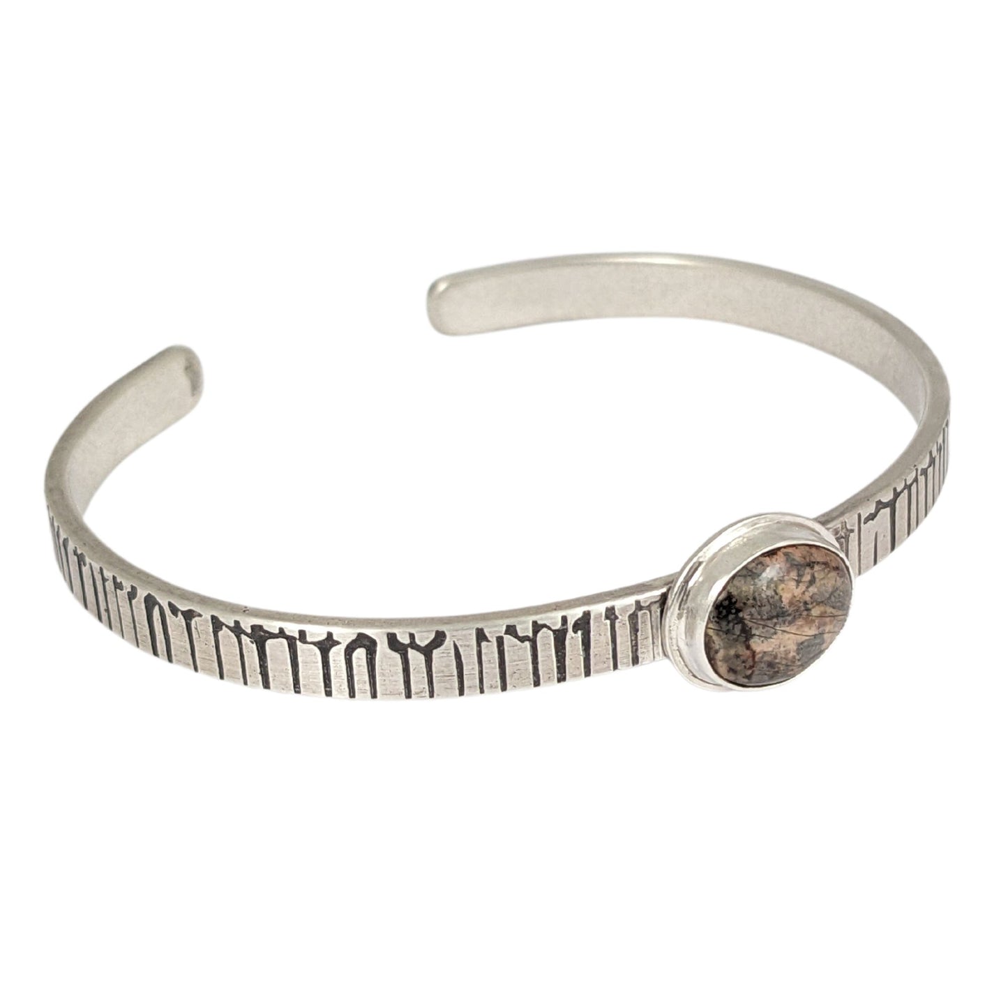 A stone in muted shades of pink, gray, and brown set on a narrow rectangular shaped sterling silver cuff bracelet. The cuff has a design that looks like an earthquake seismograph chart.