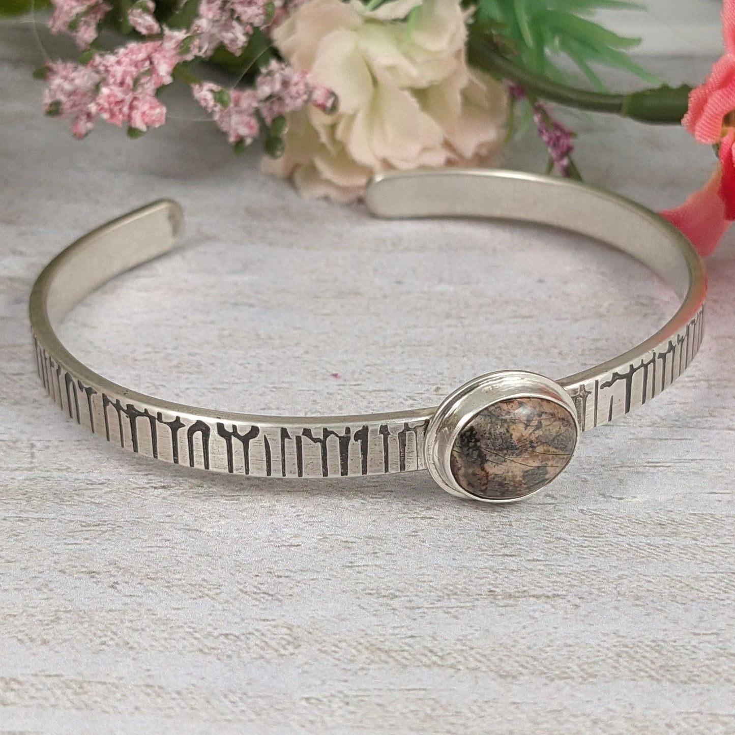A stone in muted shades of pink, gray, and brown set on a narrow rectangular shaped sterling silver cuff bracelet. The cuff has a design that looks like an earthquake seismograph chart. Staged with flowers in the background.
