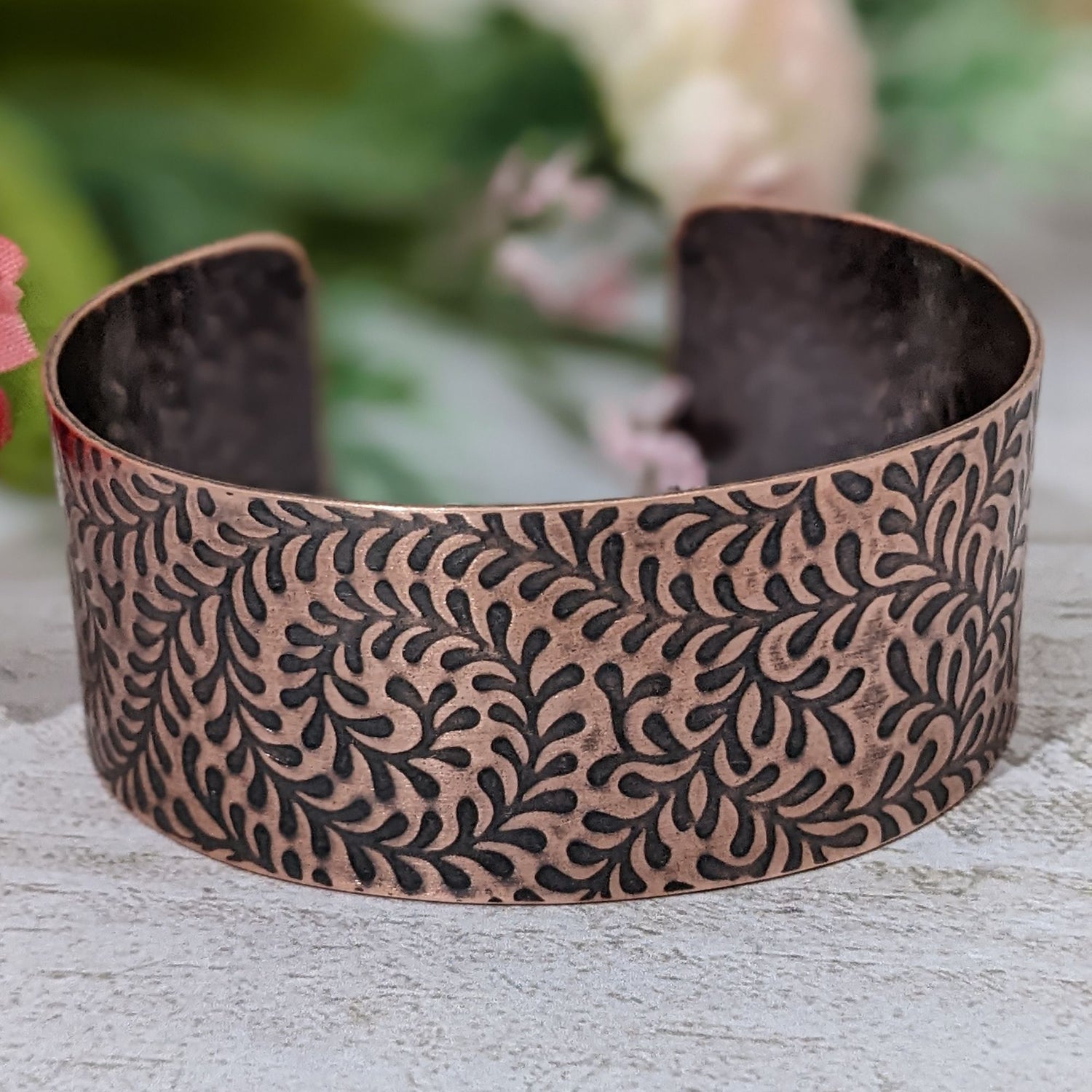 One inch wide copper cuff bracelet with impression design of a fiddlehead fern. The design is abstract. The recessed parts of the design are darkened to accentuate the detail.