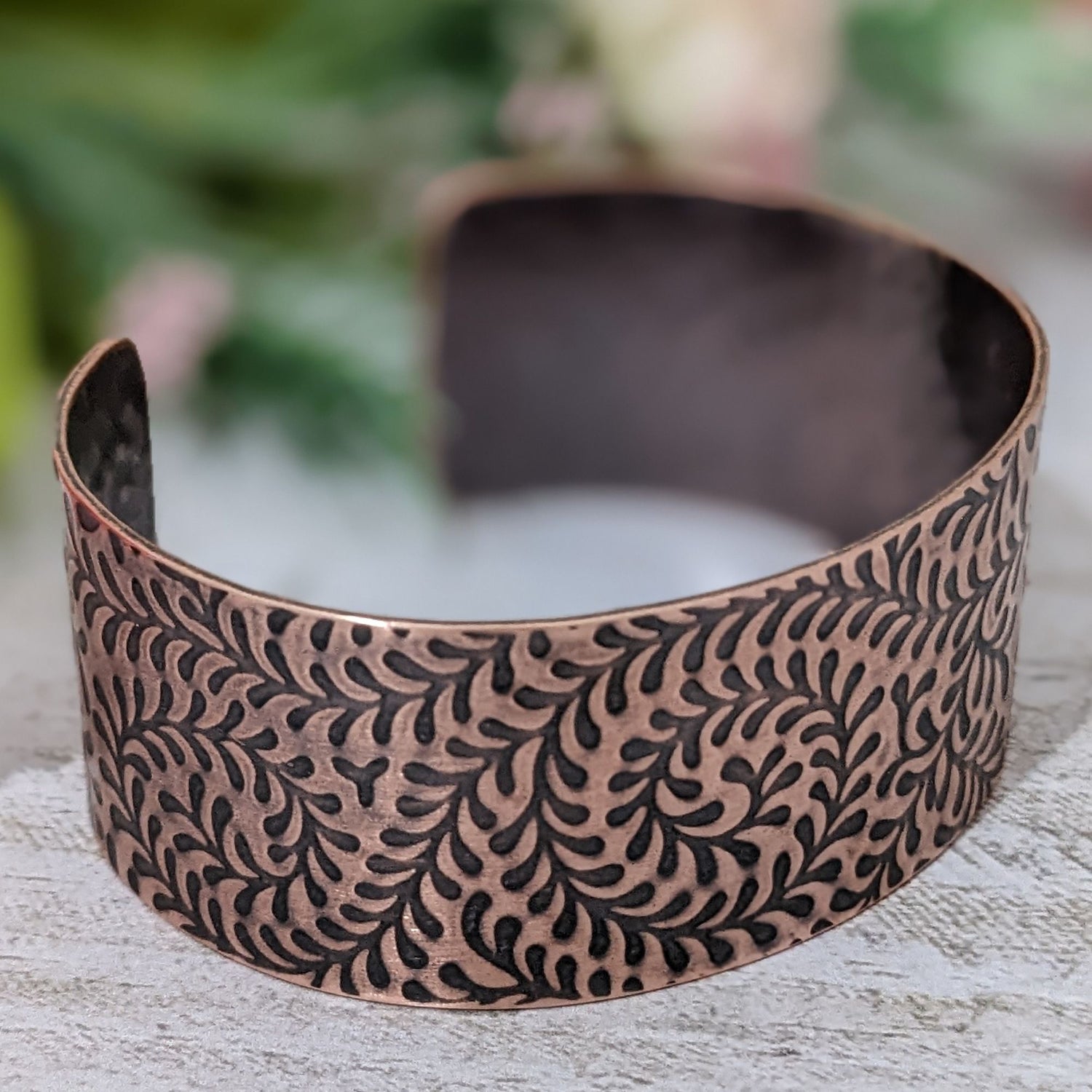 One inch wide copper cuff bracelet with impression design of a fiddlehead fern. The design is abstract. The recessed parts of the design are darkened to accentuate the detail.