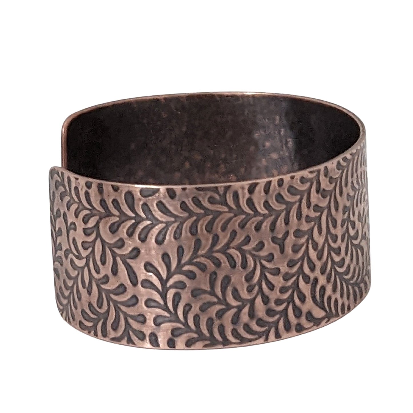 One inch wide copper cuff bracelet with impression design of a fiddlehead fern. The design is abstract. The recessed parts of the design are darkened to accentuate the detail.