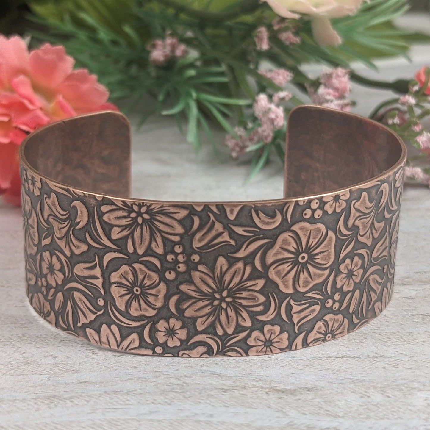 One inch wide copper cuff bracelet covered in designs of common garden flowers, looked at from above. The background around the flowers is darkened to accentuate the detail.