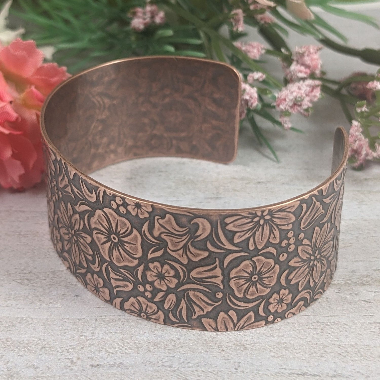 One inch wide copper cuff bracelet covered in designs of common garden flowers, looked at from above. The background around the flowers is darkened to accentuate the detail.