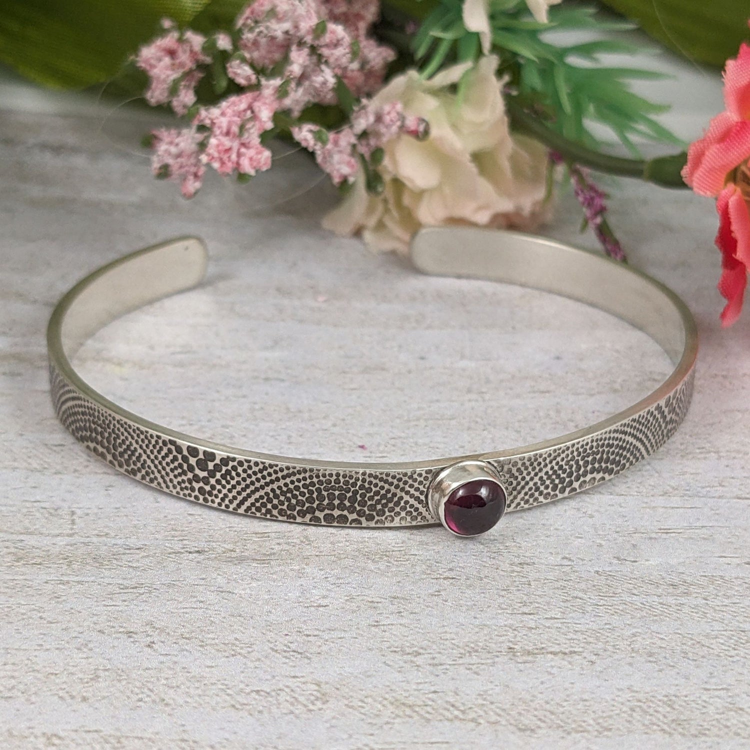 Narrow sterling silver cuff bracelet. The silver is covered with an arch pattern. In the center is a red garnet gemstone. Staged with flowers in the background.