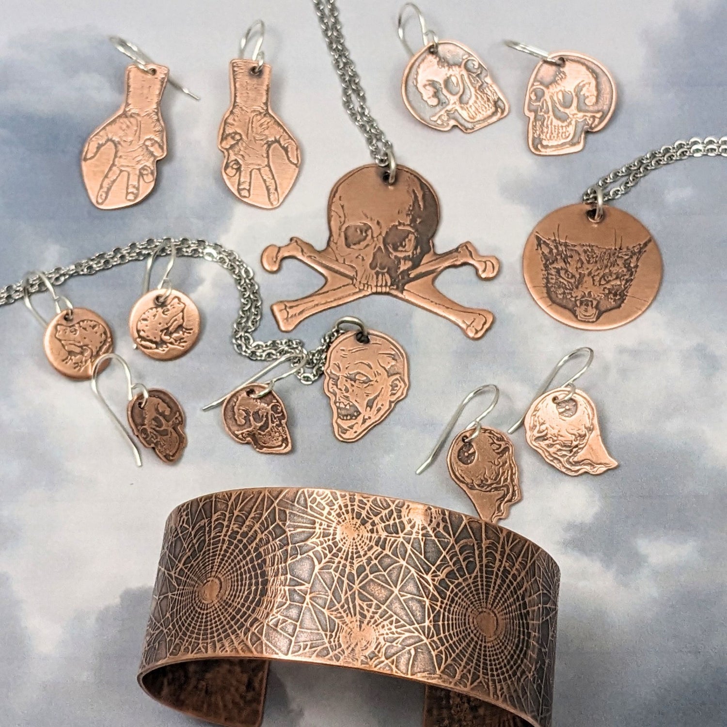 collection of halloween copper jewelry. Earrings are: hands, skulls, frogs, brains. Pendants are skull and crossbone, demon head, cat face. Cuff is spiderwebs