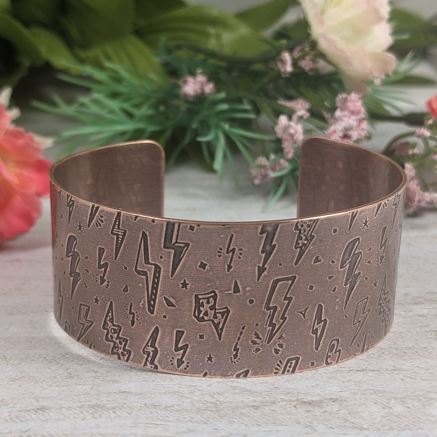 One inch wide copper cuff bracelet with an impressed pattern of assorted cartoon style lightning bolts. The bolts are darkened to accentuate the details.