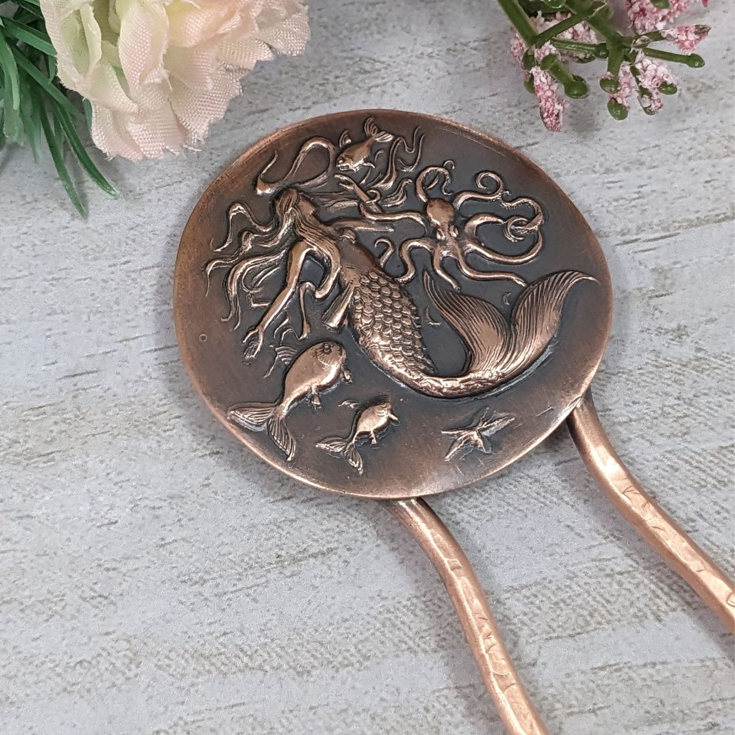 Copper hair fork. At the top is a large round disk with a sea life design impressed in the copper. There is a large mermaid with flowing hair in the center, an octopus to her left, three fish, and a starfish.