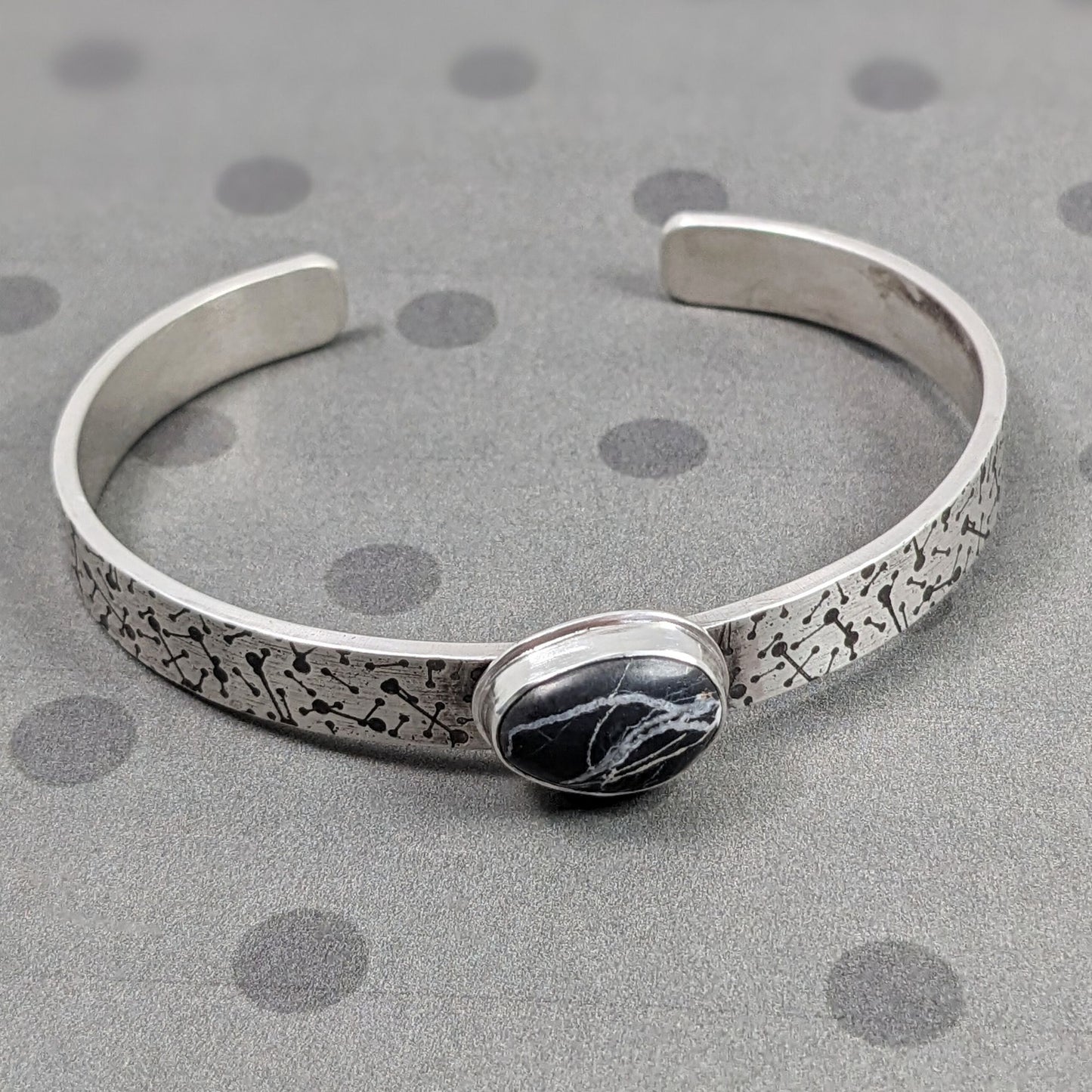 8 millimeter wide sterling silver cuff bracelet. The silver is covered in short lines with small dots on the ends that represent meteor showers. On the center is a freeform oval shaped white buffalo stone, which is a black stone with random streaks of white running through it.